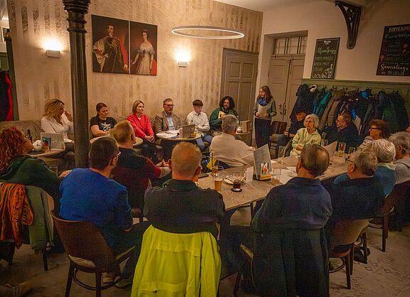Landtagskandidat*innen sitzen nebeneinander auf einer Bank im Kaffeehaus, davor aufmerksam zuhörende Menschen aus dem Publikumtgliedern und Interessierten im Kaffeehaus Gutmann