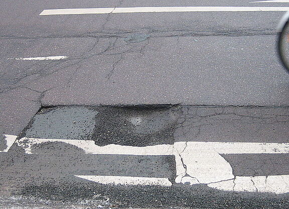 Ein Schlagloch auf der Straße, man sieht noch das Hinterrad eines vorbeifahrenden Fahrrads.