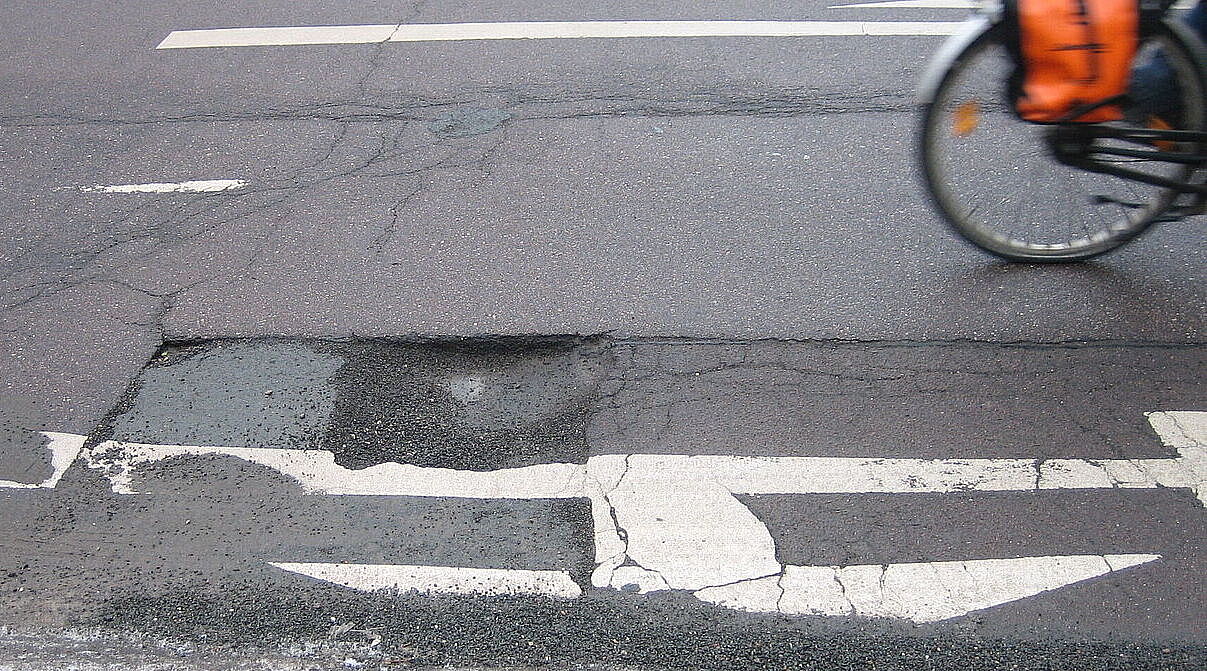 Ein Schlagloch auf der Straße, man sieht noch das Hinterrad eines vorbeifahrenden Fahrrads.