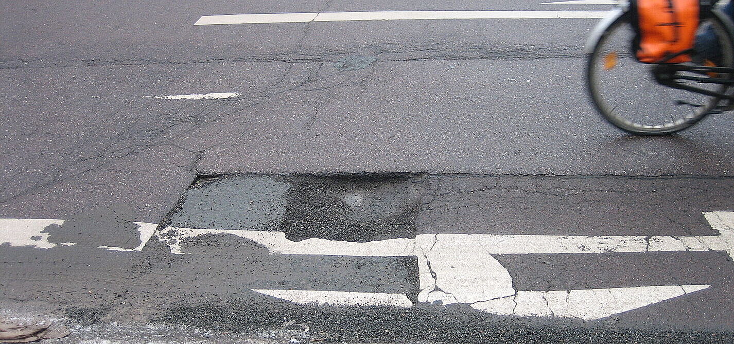 Ein Schlagloch auf der Straße, man sieht noch das Hinterrad eines vorbeifahrenden Fahrrads.